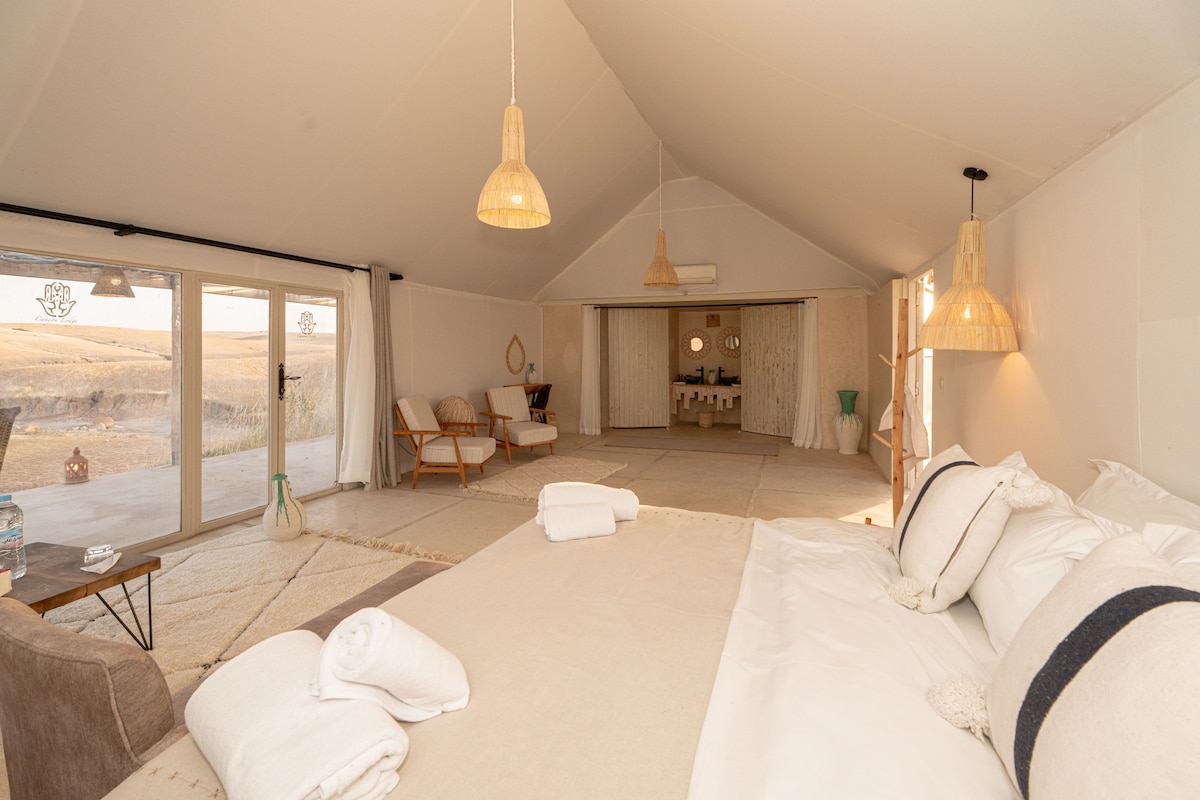 Une chambre spacieuse avec un grand lit, un coin salon avec des fauteuils et une vue sur l’extérieur, accessible par une porte coulissante.