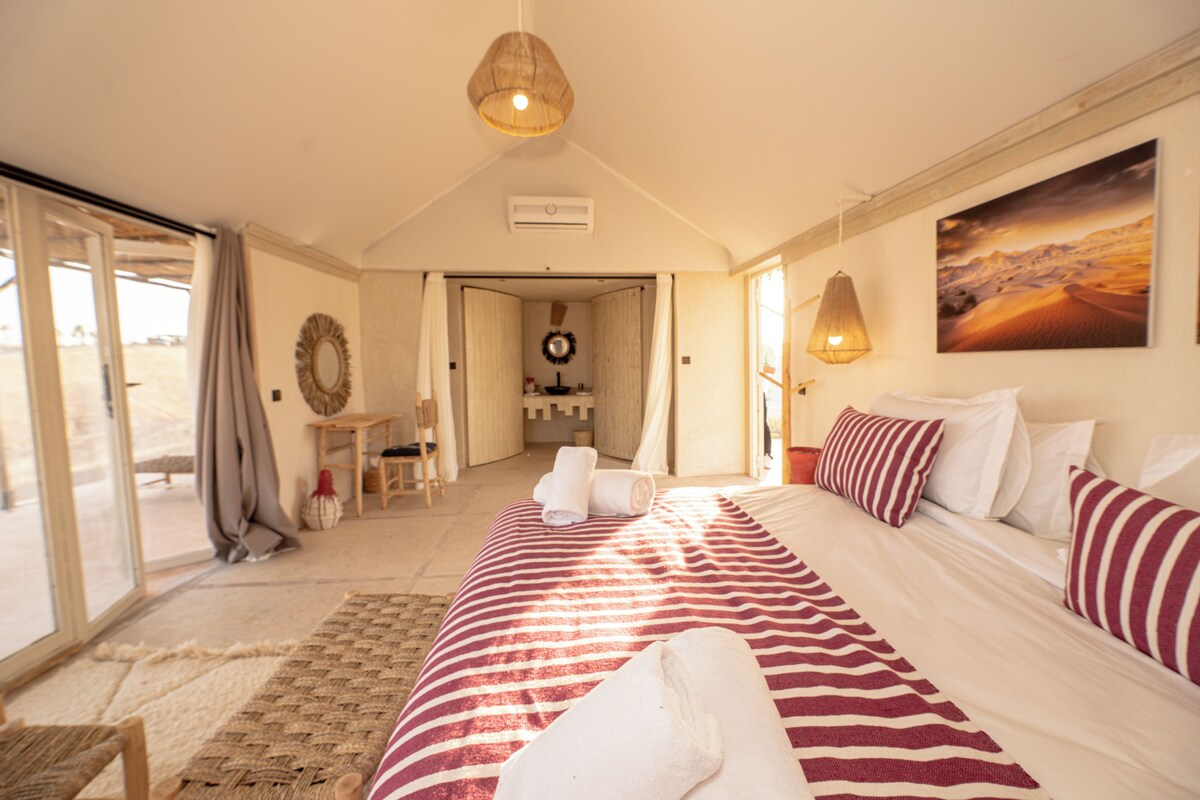 Une chambre d’hôtel avec un grand lit, une vue sur le désert par la fenêtre, une porte donnant sur un balcon et une petite table avec une chaise.
