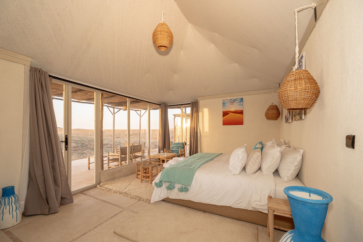Une chambre à coucher avec un lit, des oreillers, une table de chevet, une fenêtre avec des rideaux et une vue sur l’extérieur.