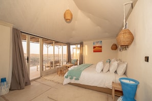 A bedroom with a bed, pillows, a bedside table, a window with curtains, and a view of the outdoors. - Mirage Agafay Luxury Camp (Sidi Badhaj)