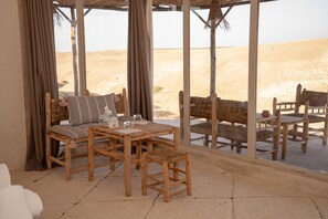 A room with a wooden table and chairs, a sofa, and a view of the desert through large windows. - Mirage Agafay Luxury Camp (Sidi Badhaj)