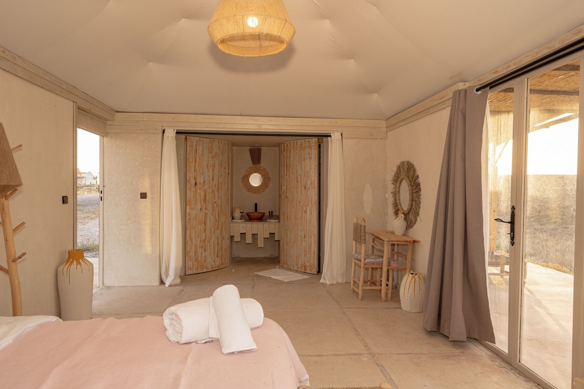 Une chambre d’hôtel comprenant un lit, un bureau, une chaise, une salle de bain avec un lavabo et un miroir, et une vue accessible par une porte coulissante.
