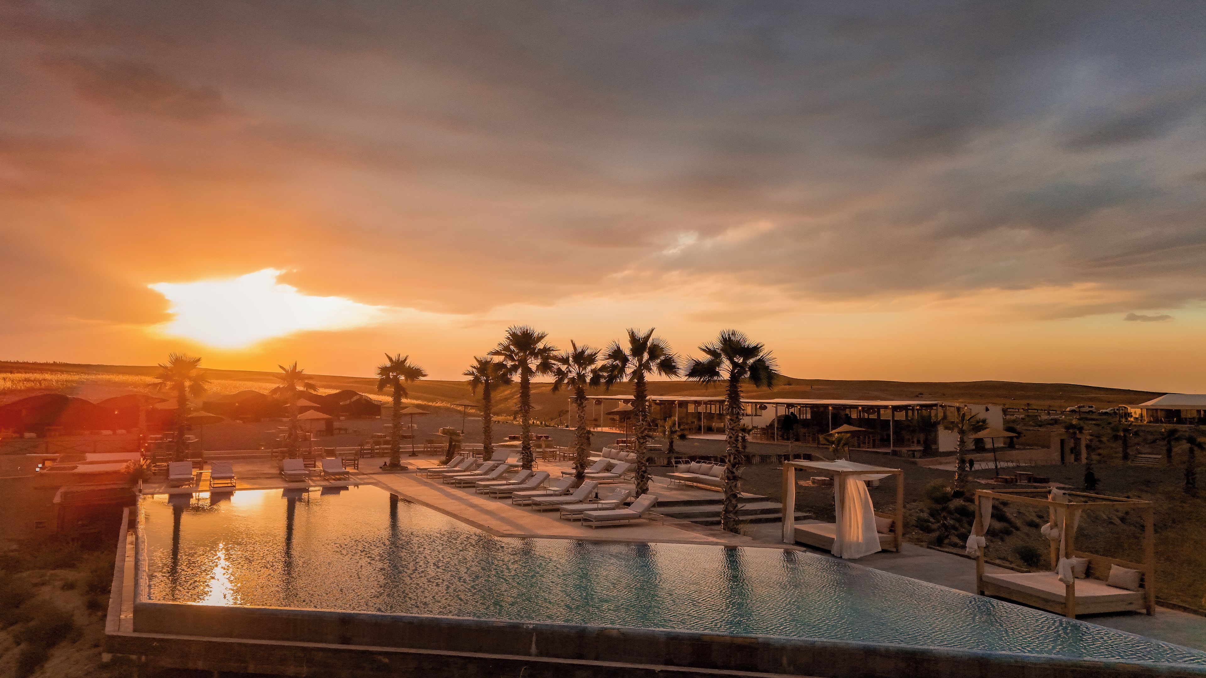 A resort pool area with lounge chairs and palm trees, surrounded by buildings and a sunset in the background.