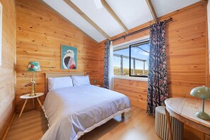 A wooden cabin bedroom with a bed, a nightstand, a lamp, a window with curtains, and a painting on the wall.