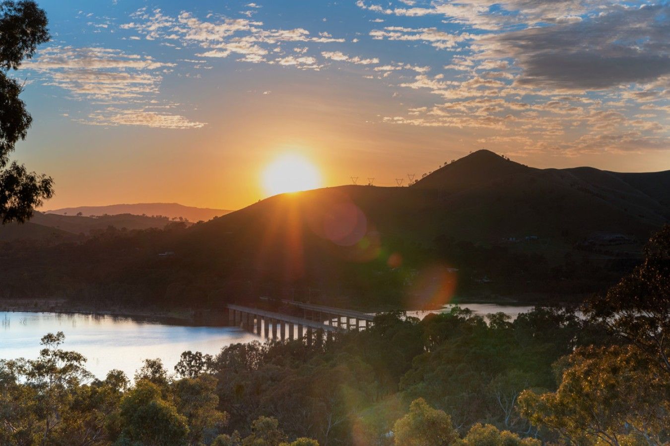 A sunset over a lake with a bridge and hills in the background.