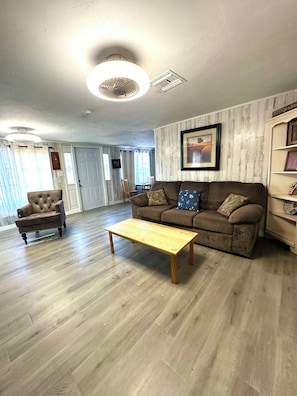 A living room with a wooden floor, a brown sofa, a wooden coffee table, and a tufted armchair. - Lovely 3-bedroom house with AC in North Fort Myers pet friendly (North Fort Myers)