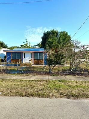 Ein einstöckiges Haus mit blauem Dach, einem eingezäunten Garten und einem kleinen Baum.