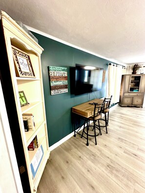 A dining area with a table and chairs, a television mounted on the wall, and a bookshelf with various items. - Lovely 3-bedroom house with AC in North Fort Myers pet friendly (North Fort Myers)