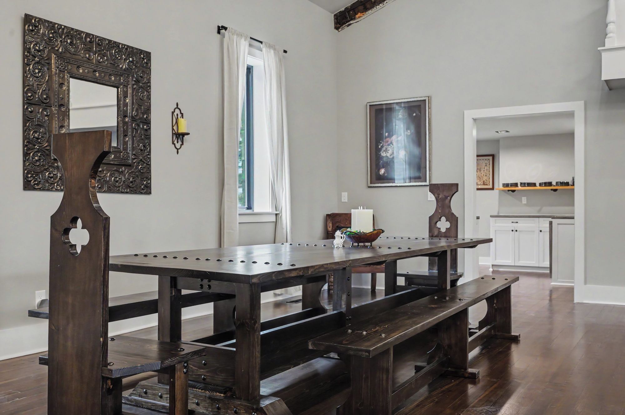 A dining area with a wooden table and benches, a decorative mirror, a wall-mounted light, and a window with curtains.