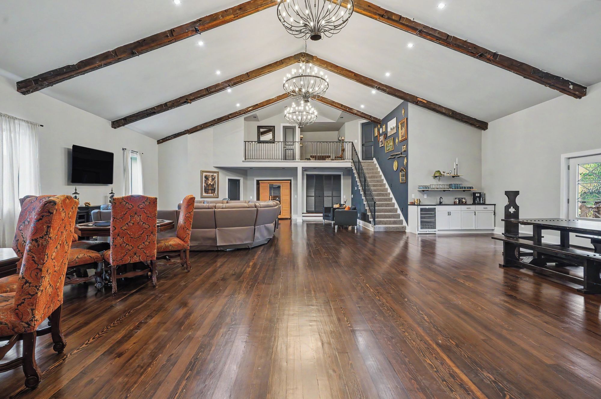 A spacious living area with a high ceiling and exposed wooden beams, a large chandelier, and a staircase leading to an upper level.