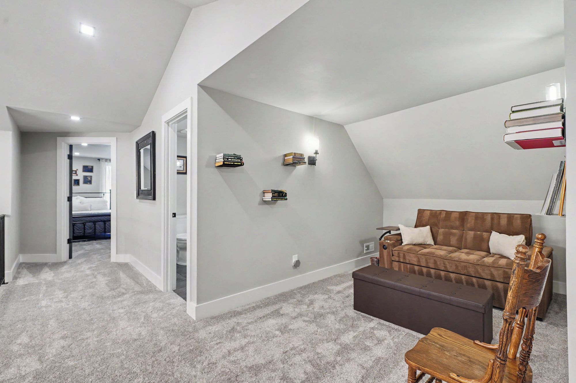 A bedroom with a sloped ceiling, a brown sofa, a wooden chair, and a bookshelf.