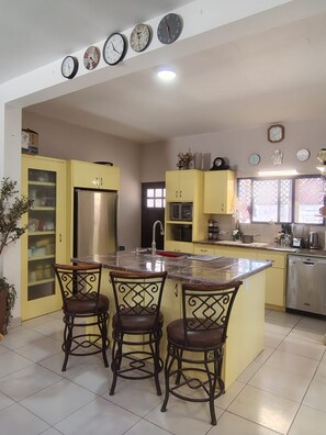 A kitchen with a central island, yellow cabinets, a refrigerator, and a sink. There are three chairs at the island and multiple clocks on the wall. - Spacious 2-Bedroom Ground-Floor Home in Downtown Loreto (Loreto)