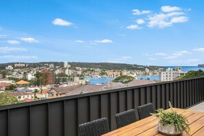 Ein Balkon mit Blick auf eine Küstenstadt, einem Holztisch und schwarzen Korbstühlen.