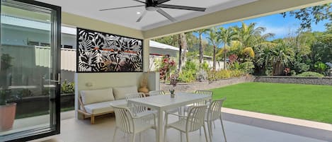 Un espacio moderno para comer al aire libre, con ventilador de techo, puerta de vidrio que da a un jardín y mesa blanca con sillas.