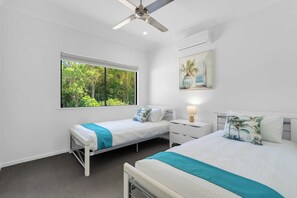 A bedroom with two single beds, a ceiling fan, a window with a view of trees, and a wall-mounted air conditioning unit. - Cedar Cove - Tropical Escape near Palm Cove Beach (Palm Cove)