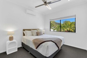 Ein Schlafzimmer mit einem Bett, einem Nachttisch, einer Lampe, einem Deckenventilator und einem Fenster mit Blick auf Grünflächen.