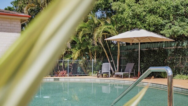 En swimmingpool med en metalstige, omgivet af et hegn, med liggestole og en parasol i et skyggefuldt område.