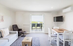 Een woonkamer met een bank, een salontafel, een aan de muur bevestigde televisie en een eethoek met stoelen en een tafel.