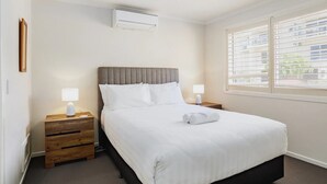 En pænt redt seng med hvidt sengetøj, to sengeborde med lamper, et vindue med persienner og en aircondition-enhed på væggen.
