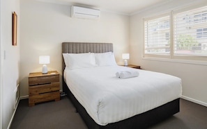 Een netjes opgemaakt bed met witte beddengoed, twee nachtkastjes met lampen, een raam met zonwering en een airconditioning aan de muur.