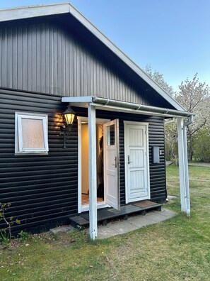 Ein kleines, eingeschossiges Gebäude mit dunkler Fassade, einer weißen Tür und einer Veranda mit einer Lampe.