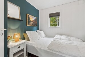 Ein Schlafzimmer mit einem Bett, einem Nachttisch mit Lampe, einem Bild an der Wand und einem Fenster mit Blick auf Bäume.