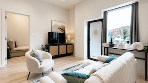 Una sala de estar moderna con un conjunto de sofás, un televisor y una ventana con vista a la calle.