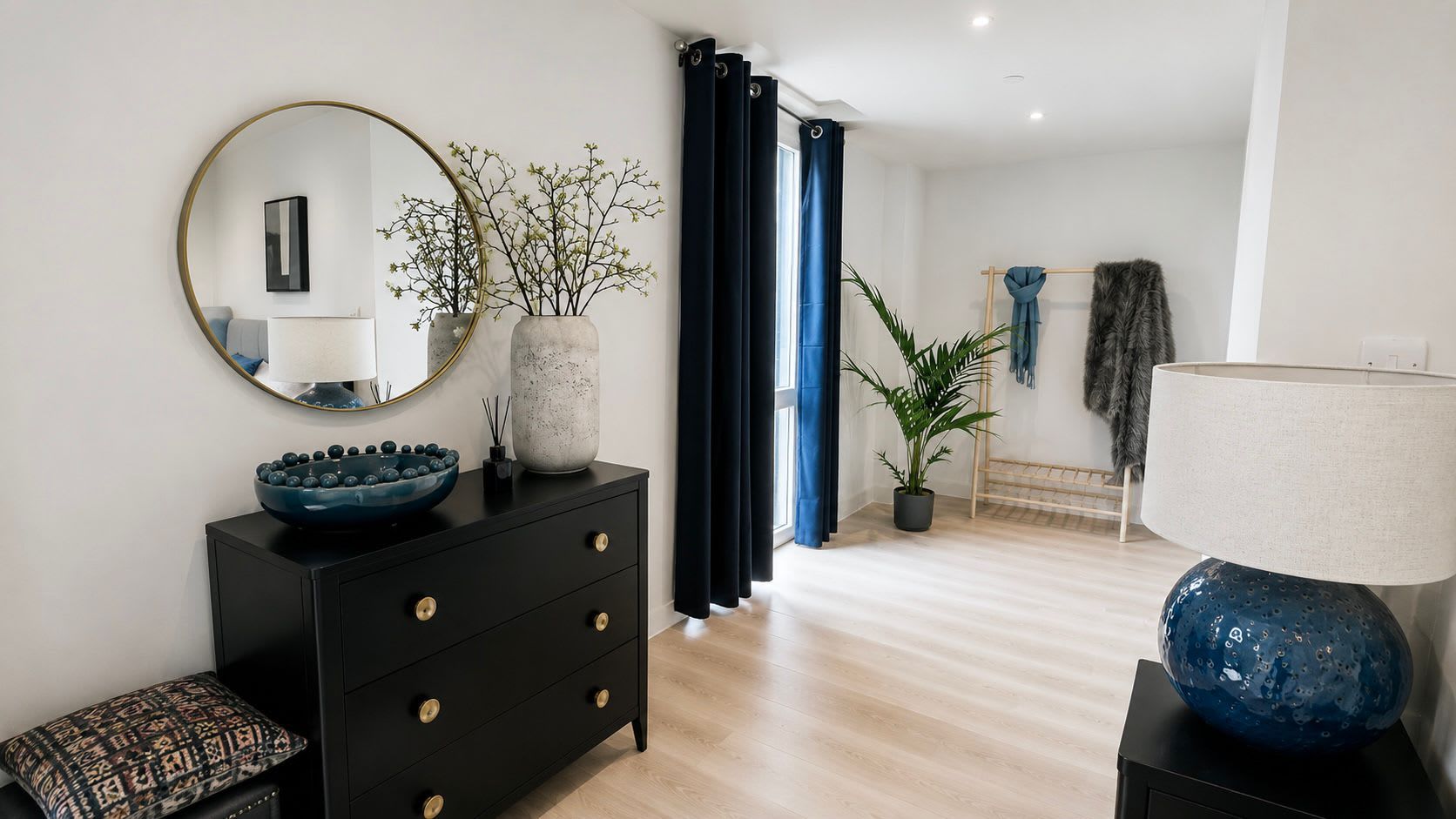 Et moderne værelse med et sort kommode, et rundt spejl, en blå skål, en potteplante og en blå lampe.