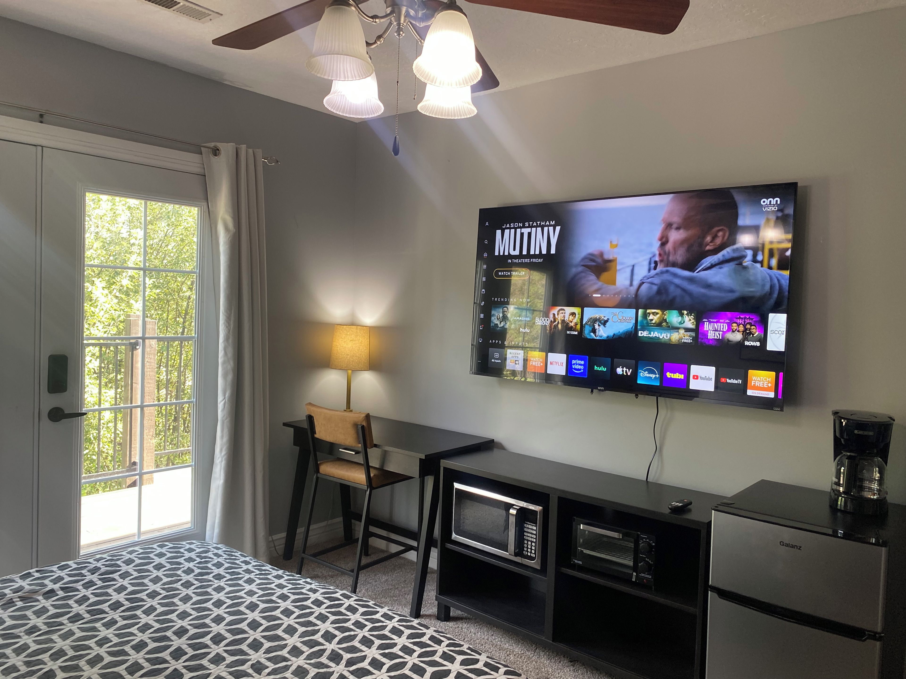 A hotel room with a bed, a desk with a lamp, a television mounted on the wall, and a minibar.