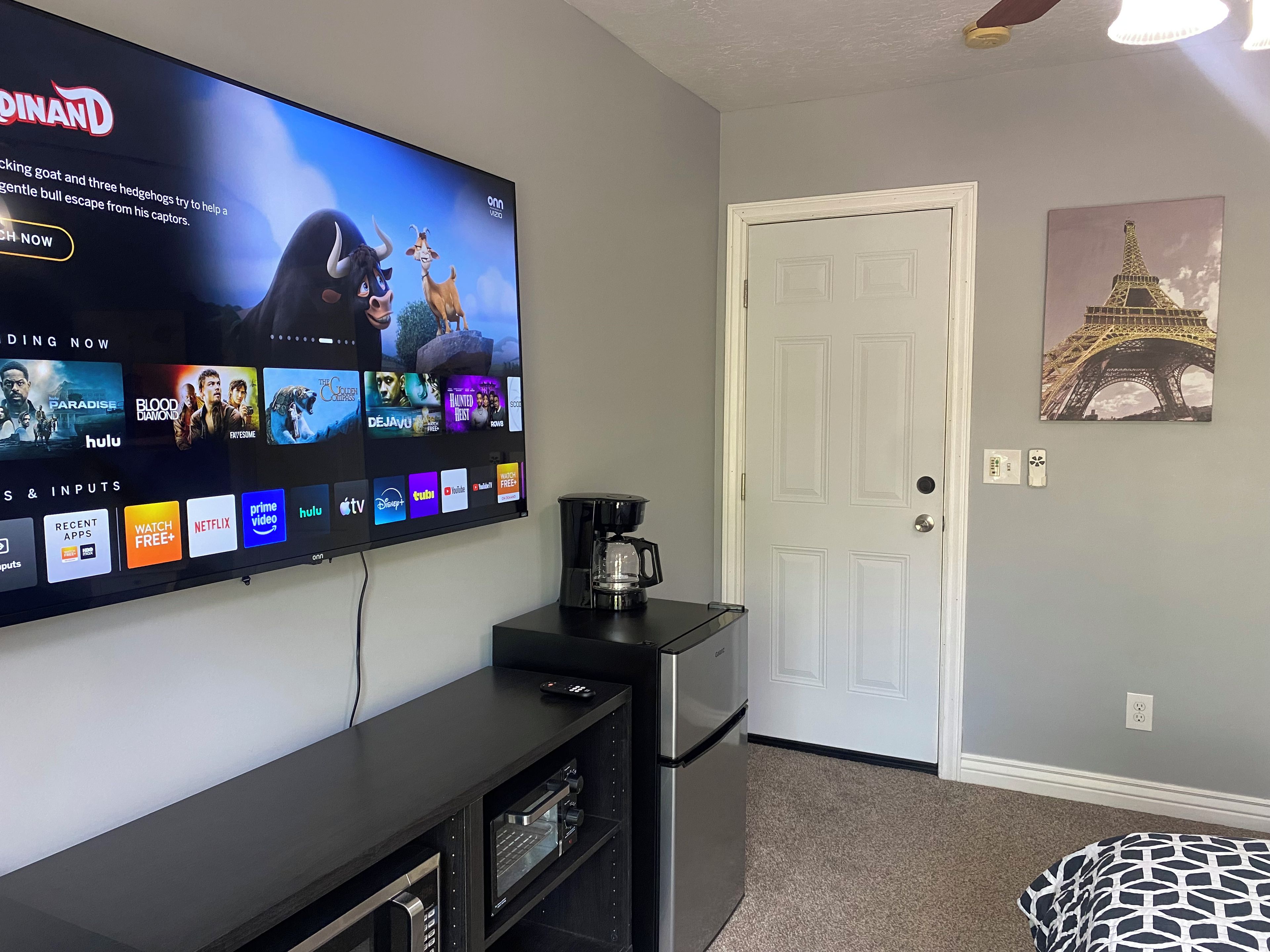 Ein Wohnzimmer mit einem Flachbildfernseher, der eine Streaming-Dienstanzeige zeigt, einer Kaffeemaschine, einer Tür und einem Wandbild der Eiffelturms.