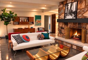 A modern living room with a stone fireplace, a white sofa with patterned cushions, a glass coffee table, and a large plant. - Custom Mid Century! Game Room, Sleeps 15.  4 King Rooms. Theatre Room. (Mesa)