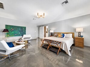 Ein modernes Hotelzimmer mit einem großen Bett, einer Sitzecke mit Sessel und kleinem Tisch, einem Bild an der Wand und einer Deckenleuchte.
