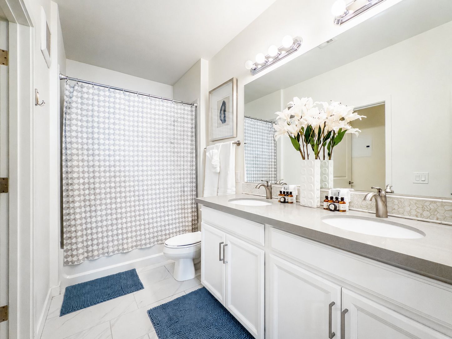 A bathroom with a shower, toilet, two sinks, a mirror, and a vase with flowers.