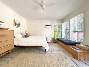 Une chambre à coucher comprenant un lit, une commode, une table de chevet, une lampe, un appareil de climatisation, une fenêtre avec des stores et une vue sur la verdure à l’extérieur.