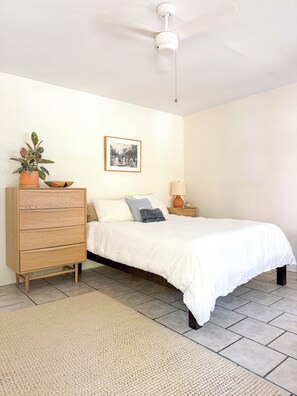 Een slaapkamer met een bed, een houten dressoir, een nachtkastje, een lamp, een plafondventilator en een ingelijst schilderij aan de muur.