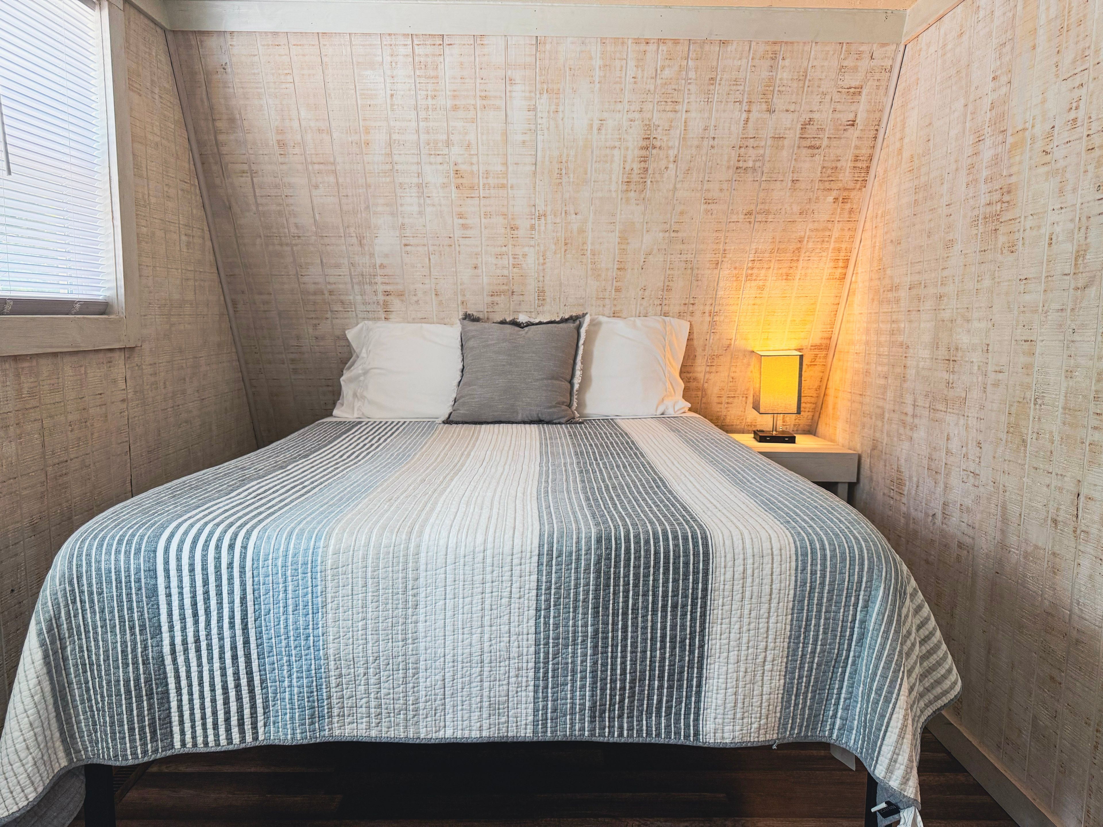Ein Schlafzimmer mit schrägen Wänden, einem Bett mit gestreifter Bettdecke, zwei Kissen, einem Nachttisch mit Lampe und einem Fenster mit Jalousien.