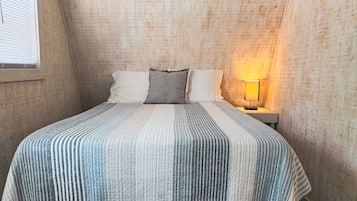 Ein Schlafzimmer mit schrägen Wänden, einem Bett mit gestreifter Bettdecke, zwei Kissen, einem Nachttisch mit Lampe und einem Fenster mit Jalousien.