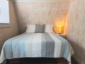 Een slaapkamer met een schuine kap, een bed met een gestreept dekbedovertrek, twee kussens, een nachtkastje met een lamp en een raam met jaloezieën.