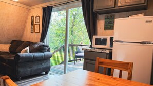 Een gezellige leefruimte met een bank, een houten tafel, een keuken met een koelkast en magnetron, en een schuifdeur die uitkomt op een terras.