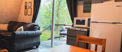 Een gezellige leefruimte met een bank, een houten tafel, een keuken met een koelkast en magnetron, en een schuifdeur die uitkomt op een terras.