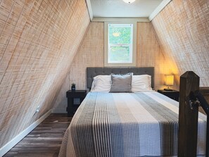 A bedroom with a sloped ceiling, a bed with a grey headboard, a nightstand with a lamp, and a window with a view of greenery. - Table Rock Lake A-Frame Cabin | Black Oak Lodge 3 (Lampe)