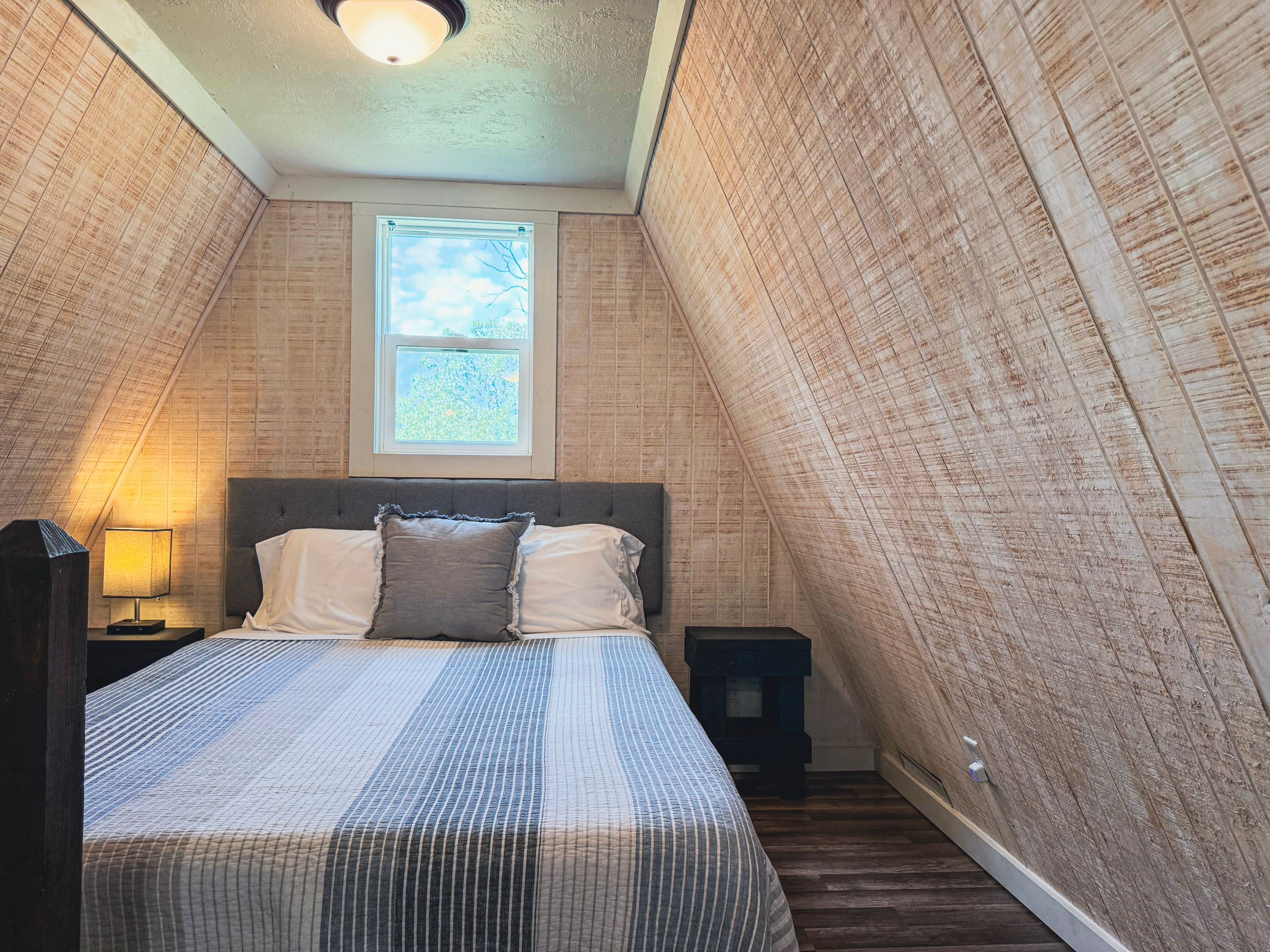 A bedroom with a sloped ceiling, a window, a bed with pillows, a nightstand, and a lamp.