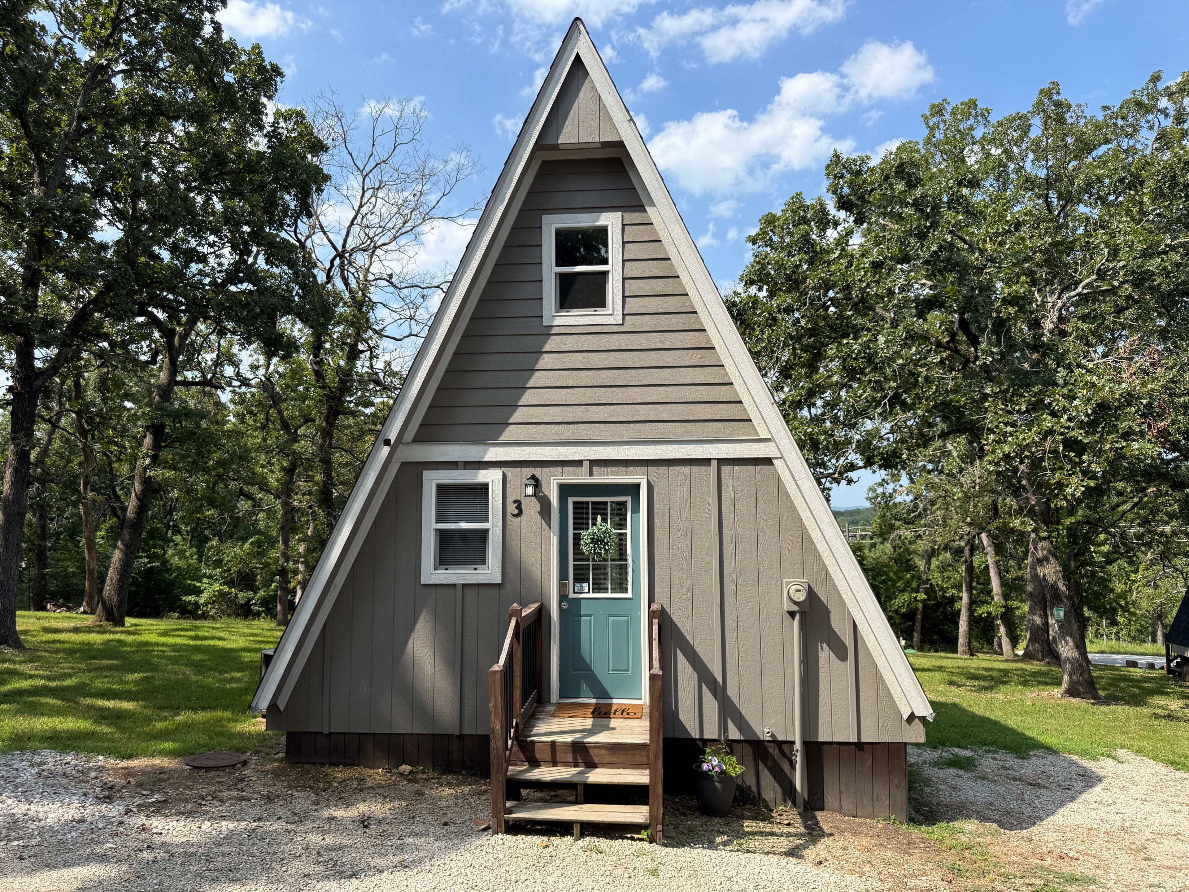 A small, triangular wooden cabin with a door and windows, surrounded by trees.