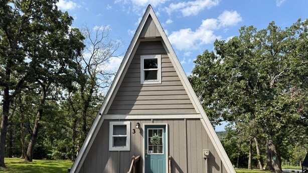 Een klein, driehoekig houten huisje met een voordeur en een klein terras.
