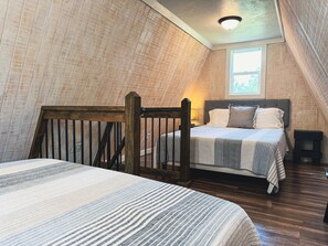 Un dormitorio con techo inclinado, una cama con cabecera, una silla de madera, una mesita y una ventana.