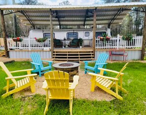 Een overdekt buitengebied met Adirondack-stoelen en een vuurkuil, omringd door een wit hek en een camper.