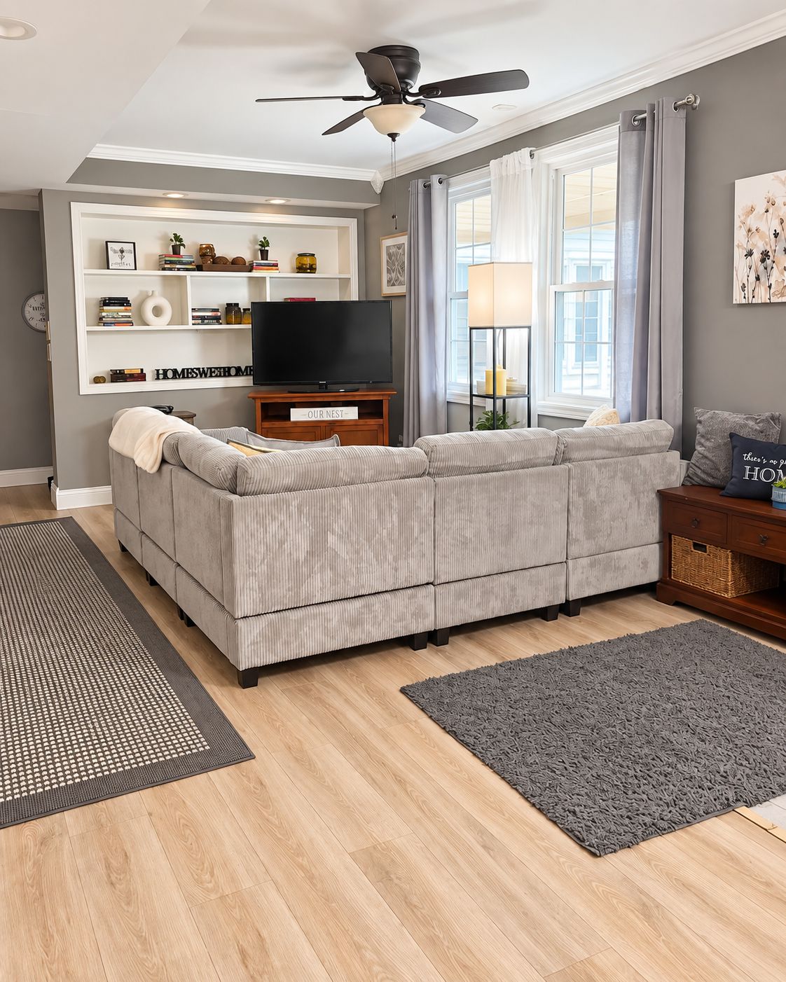 Ein Wohnzimmer mit grauem Sofa, einem Flachbildfernseher, einem Deckenventilator und einem Bücherregal mit Dekorationselementen.