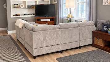 Ein Wohnzimmer mit grauem Sofa, einem Flachbildfernseher, einem Deckenventilator und einem Bücherregal mit Dekorationselementen.