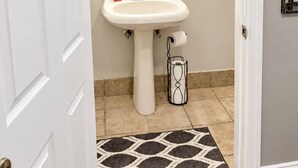 Une salle de bain avec un lavabo sur pied blanc, un miroir doté de trois points lumineux et un tapis à motifs sur le sol.