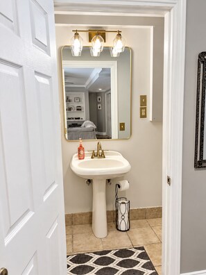 A bathroom with a white pedestal sink, a mirror with three light fixtures, and a patterned rug on the floor. - Cozy & Quiet Country 1-Bedroom Stay with Private Living Room, Bath and Entrance (Elbridge)